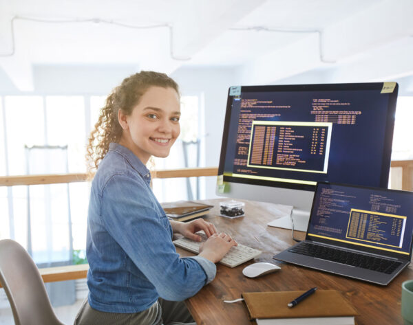 Portrait of female IT developer smiling at camera while typing on keyboard with black and orange programming code on computer screen and laptop in contemporary office interior, copy space